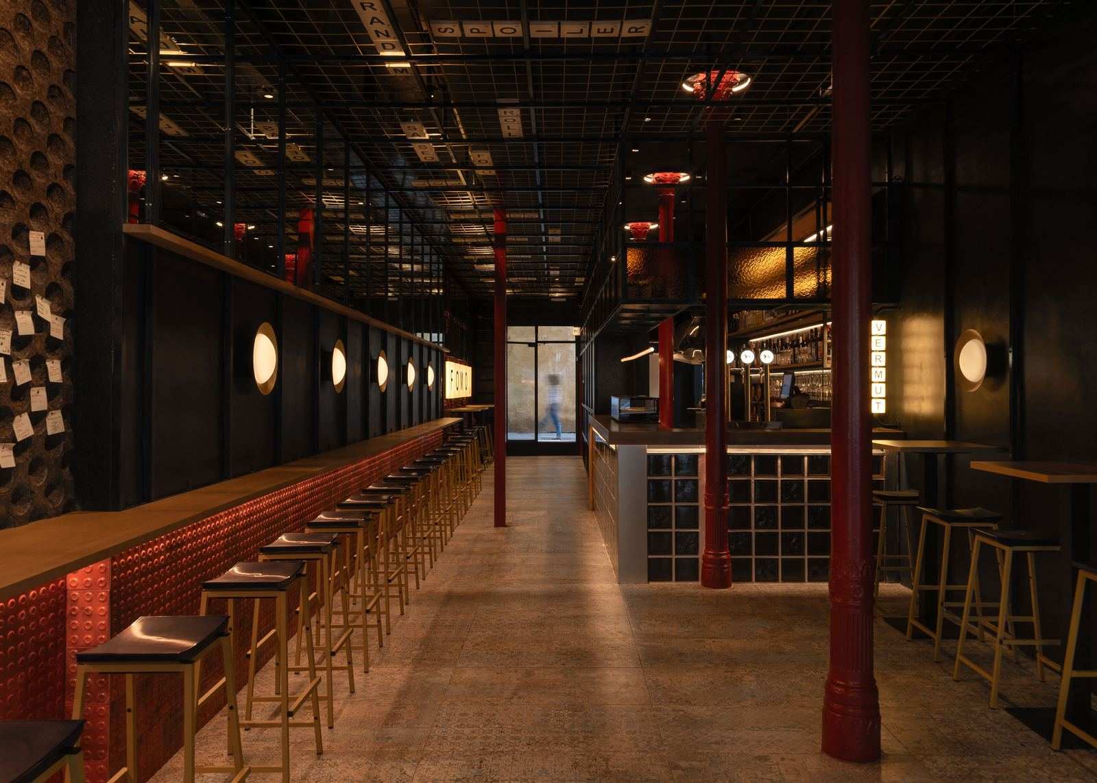 La barra de FOMO Bar: apliques redondos encendidos, frente de azulejo rojo, columnas rojas y la caja de luz VERMUT al fondo.