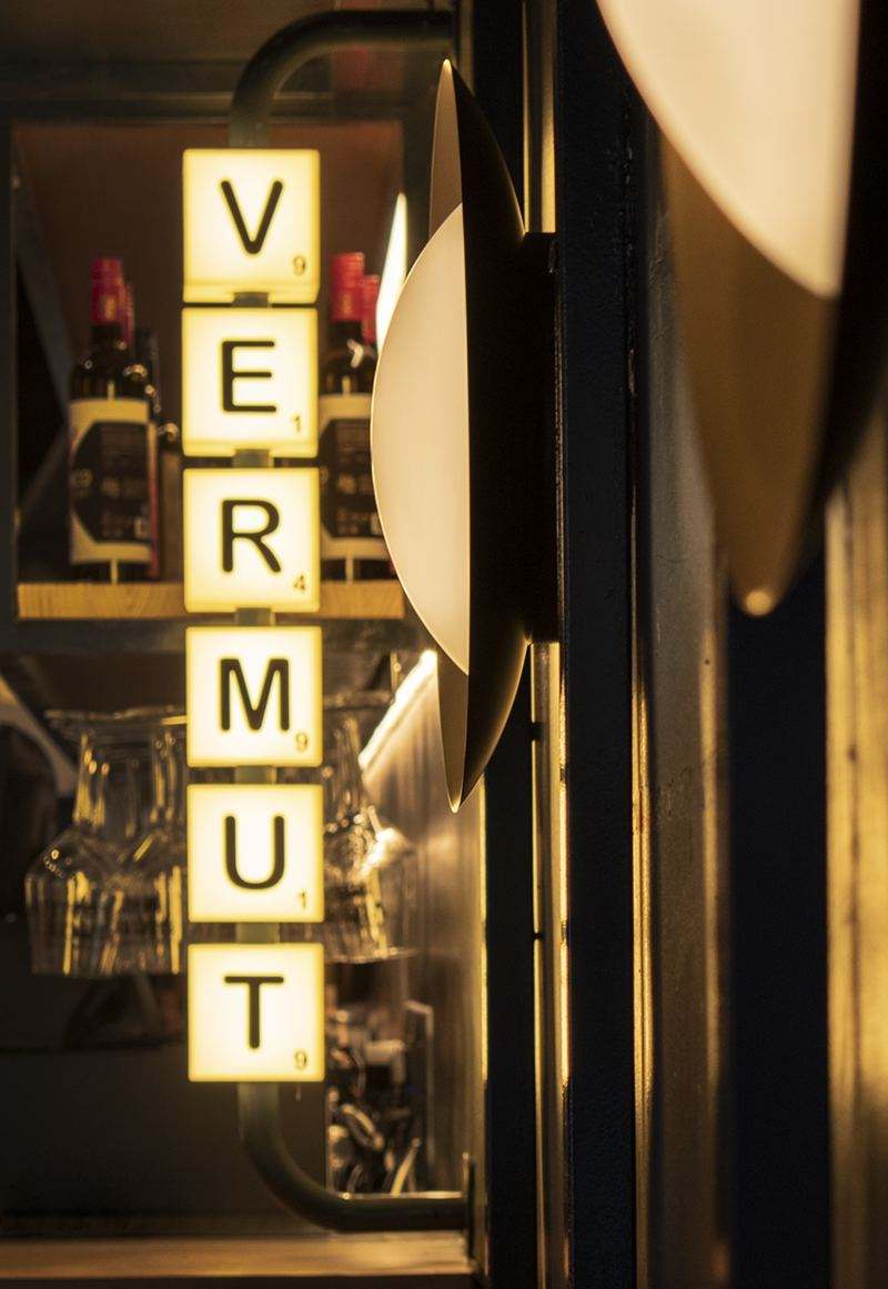 Caja de luz vertical con la palabra VERMUT en fichas iluminadas, junto a un aplique redondo y la estantería de botellas.