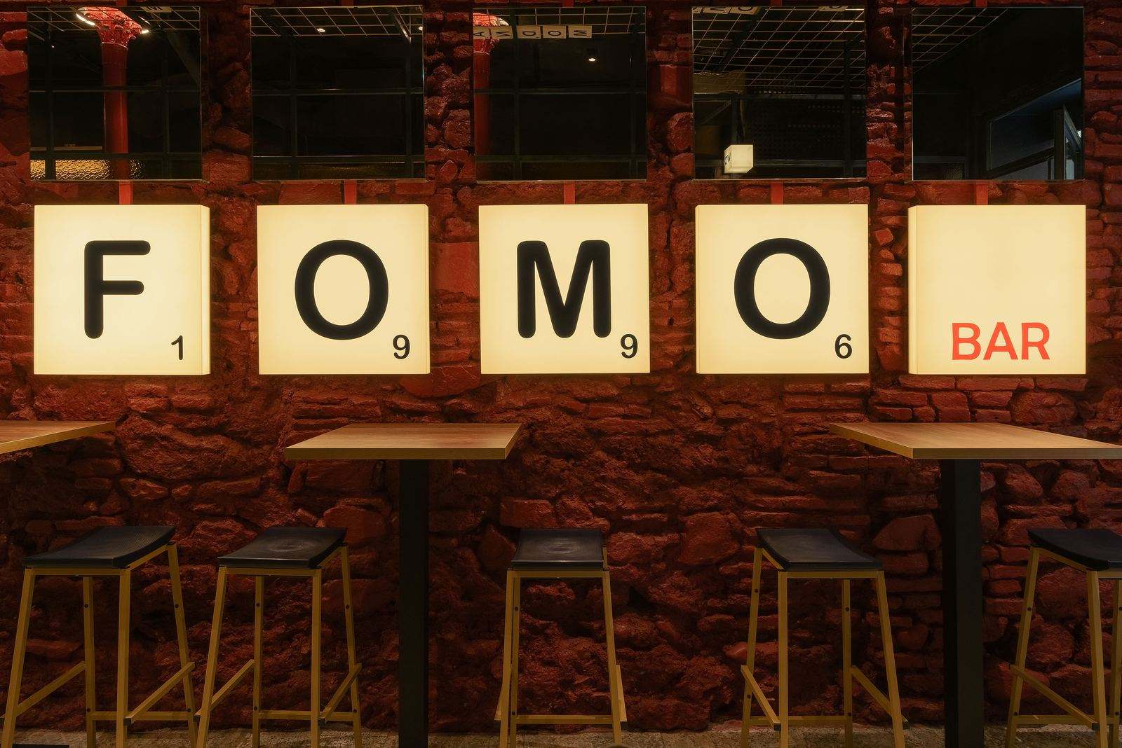 Sala de FOMO Bar: muro de piedra roja con las cajas de luz F, O, M, O y BAR sobre mesas altas y taburetes.