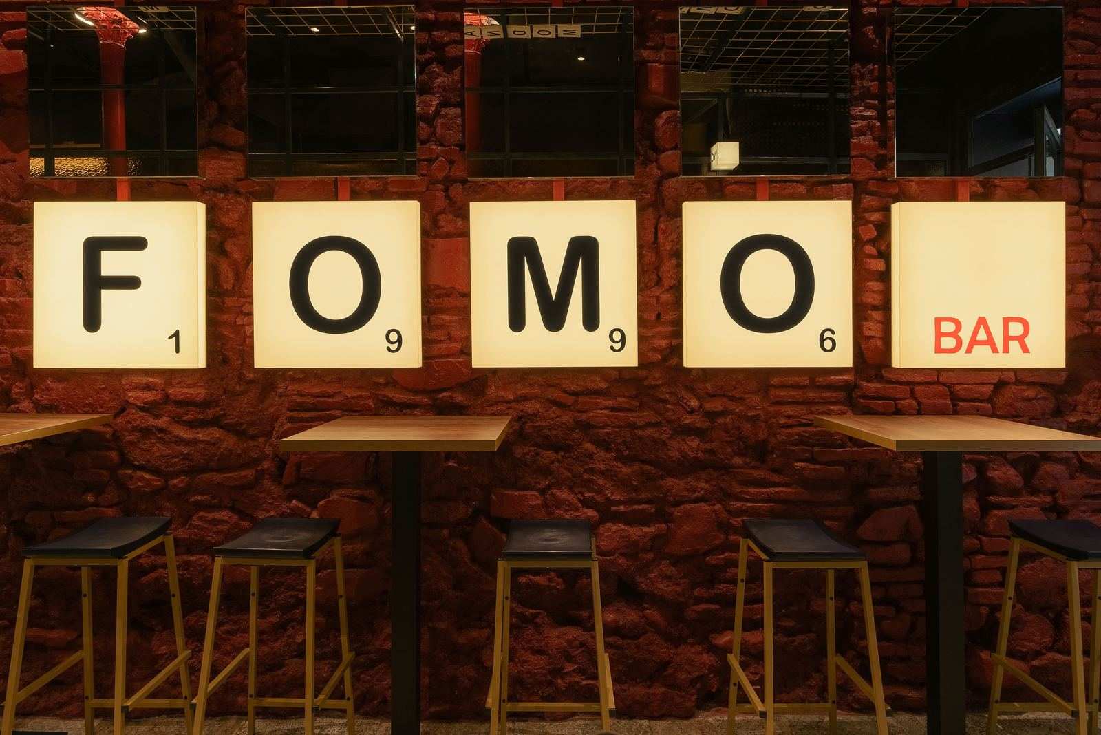 Sala de FOMO Bar: muro de piedra roja con las cajas de luz F, O, M, O y BAR sobre mesas altas y taburetes.
