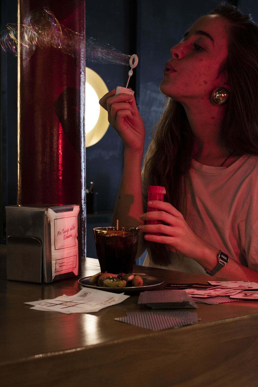 En la barra, una chica hace pompas de jabón junto a un servilletero con la servilleta «No tengo saldo. Llámame», un Duralex, un platito de aceitunas, una servilleta con un tres en raya y naipes.