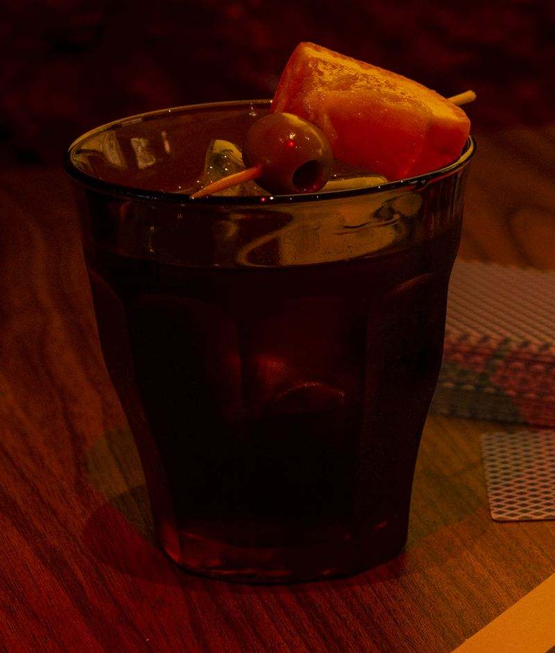 Vermut en vaso Duralex con hielo, naranja y una aceituna pinchada, sobre la mesa de madera.