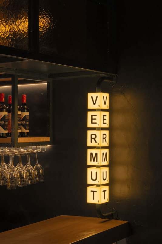 Rótulo vertical VERMUT en fichas iluminadas junto a la estantería de botellas y las copas colgadas.
