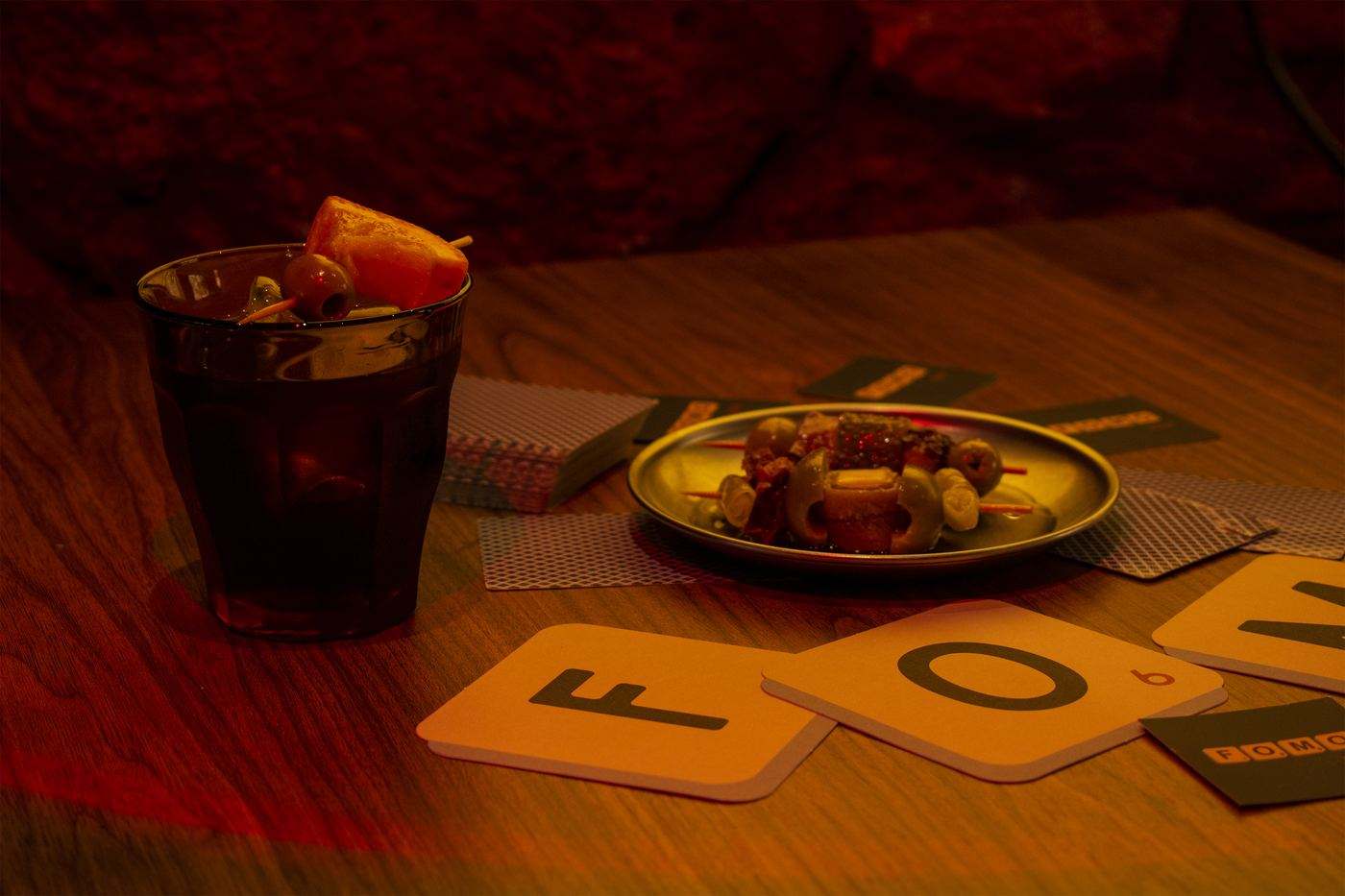 Vermut con naranja y un platito de aperitivo sobre una mesa de madera, junto a posavasos con forma de ficha F y O.